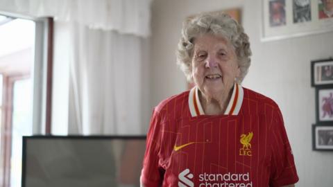 Peggy Pickavant is smiling at the camera. She is wearing a Liverpool FC shirt and is standing in her living room in front of a TV with photographs hanging on the walls.
