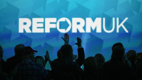 A Reform UK slogan projected on a wall with crowds pictured below in the dark.