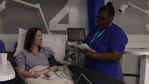 A female patient in a hospital bed is attended by a female nurse who is writing on a sheet of paper. A hospital monitor is checking the patient's blood and heart function.