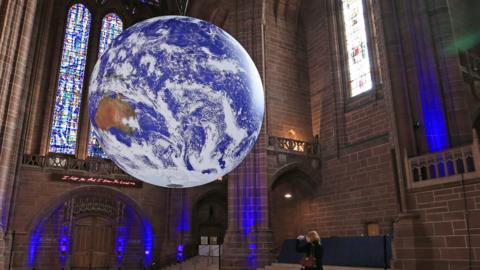 In the centre of the image is a giant scuplture of the earth called Gaia, covered in satellite imagery of the planet. It's being suspended from the ceiling. It's being suspended in Liverpool Cathedral - and globe is surrounded by stained glass window, tall brick walls, and a staircase to the right.