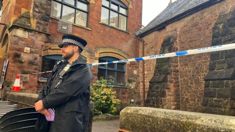 A male police officer wearing a hat and black coat stood in front of blue and white police tape. The officer is also holding a purple notebook in his left hand. The tape is surrounding a brown building on Catherine Street in Exeter. 