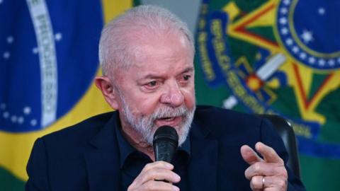 A head and shoulders shot of President Lula speaking into a microphone, with the Brazilian flag behind him