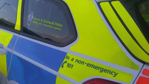The side of a yellow and blue police vehicle. On the back window, there are the words 'Devon and Cornwall Police' in yellow writing with a badge to the left of it.  On the back of the police car is the words 'In a non-emergency' call 101. 