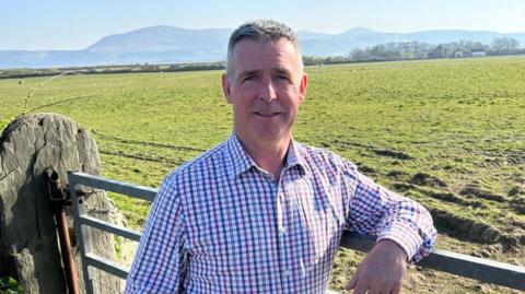John Teare has short grey hair, and wears a blue and red checked shirt. He leans on a farm gate near a green field.