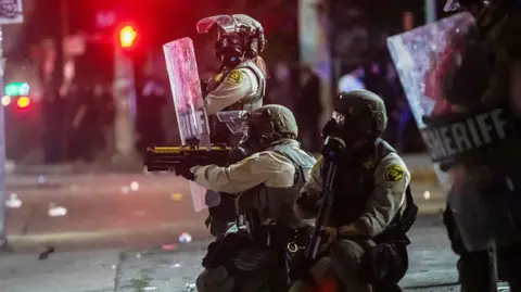 Members of the LA County Sheriff's Department in riot gear, two crouched to the ground holding guns, one female one standing holding a shield