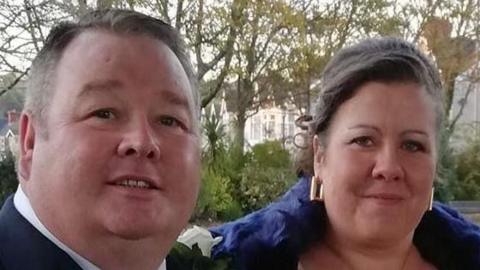 Christopher Mills a Michelle Mills ar ddiwrnod eu priodas. Christopher Mills mewn siwt ddu a Michelle Mills yn gwisgo sgarff las a chlustlysau aur.