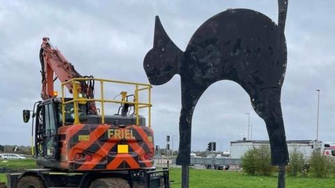 A sculpture of a black cat on the right, with its back arched and tail raised at the Black Cat roundabout, Cambridgeshire. On the left is a vehicle with an orange crane.