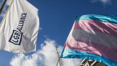 An LGB Alliance Conference flag with a transgender pride flag. Both flying in the wind.