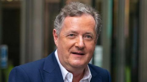 Broadcaster Piers Morgan. His head and shoulders are seen as he walks in front of a set of revolving doors. He is wearing a blue suit.