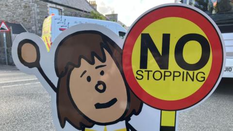 A Parking Buddy sign. It is a cartoon child with brown hair holding a yellow and red 'no stopping' sign. The sign is on the road outside a school and a white bus and white van are behind the sign.