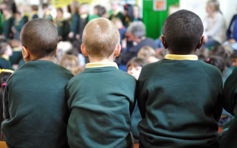 The image shows three children sitting side by side, viewed from behind. They are wearing matching dark green sweaters with yellow collars, suggesting a school uniform. The background is slightly out of focus, but it looks like an indoor space with bright colours, including a green wall or partition.