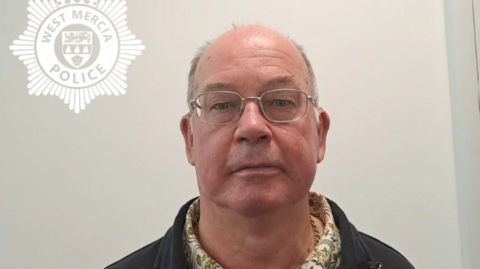 Martin Cowley pictured looking directly at the camera. He is partially bald and clean shaved. He has thin silver glasses and is wearing a floral patterned shirt and black jacket.