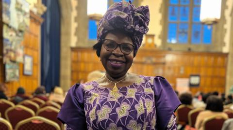 A woman in her 60s stands smiling in front of rows of seats in an opulent event room. She is wearing a traditional Nigerian dress and headpiece coloured purple, lilac and gold.