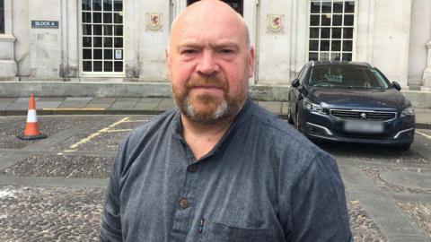 Bill Revans standing on a cobbled area outside the County Hall building in Taunton. Car parking spaces and a car are visible behind him. He has a beard and is wearing a dark blue shirt with the sleeves rolled up to his elbows.
