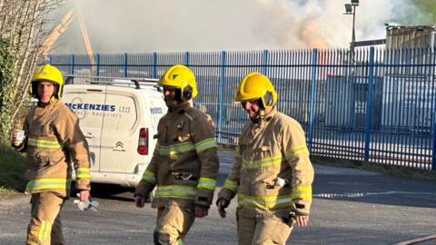 Three firefighters wearing full protective gear and bright yellow helmets walk along a road near an industrial site. Fire hoses run along the ground beside them, and thick smoke is rising in the background behind a metal fence.