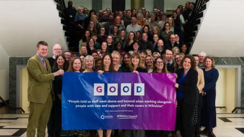 A large group of people stood on a stairway smiling. Those in the front of the group are holding an ombre blue and purple banner which reads 'GOOD' in white boxes. There is a quote at the centre of the banner which reads 'People told us staff went above and beyond when working alongside people with care and support and their carers in Wiltshire.' The CQC logo is below it, as is the logo for Wiltshire Council.