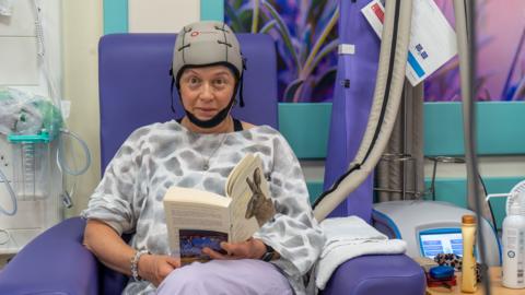 A woman in a hospital chair in a hospital ward wearing a scalp cooling machine on her head which is grey with a black chin strap. She is wearing a grey and white blouse and reading a book.