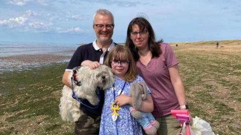 A young girl in a blue dress and blue glasses is standing with a woman in a light red/pink t-shirt and a man in a white collared navy t-shirt. They are on a beach with green seaweed on. 