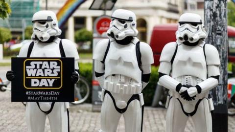 Three people dressed as Star Wars' stormtroopers hold a sign advertising Star Wars day