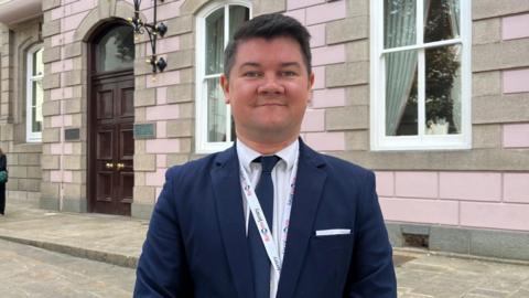 Deputy Sam Mézec is wearing a dark blue suit and is stood outside the States Assembly.