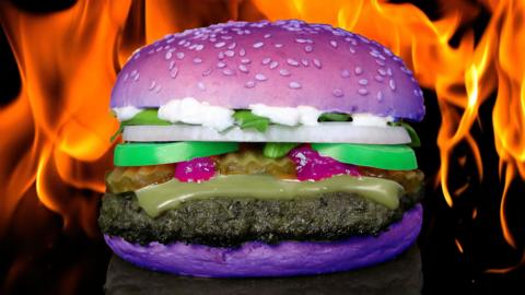 A burger feturing purple bread, green tomatoes and cheese in front of flames 