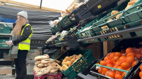 A woman in high-vis is stood in a sheltered food bank by crates of fruits and vegeatables.
