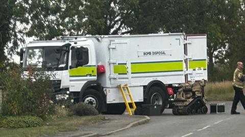 White truck with yellow and blue stripe. Bomb disposal written on it. There is a yellow ladder, a bomb disposal robot, a police car and a man in police bomb disposal officer. There are trees, bushes, a road and a path.