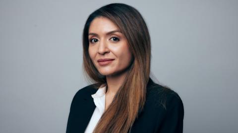 A photo portrait of Shahrzad. She has long brown hair, brown eyes, and wears a dark suit jacket and a white collared shirt.