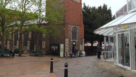 A church stands in a square, with has a shopping centre on the right. Outside the church, which is on the left, there are large trees which have circular benches around their trunks.