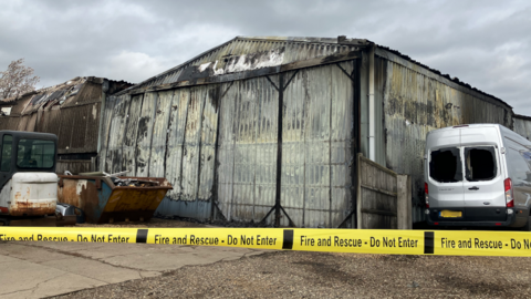 Two corrugated iron business units are charred and bent. There is a van parked next to them with smashed rear windows.