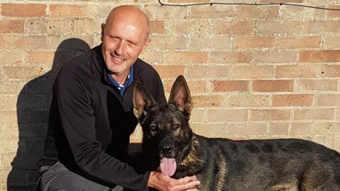 Ian Partington kneeling, holding a dark brown German Shepherd, which has its tongue out. Ian, who is bald, is wearing all black and smiling. They are in front of a brick wall.