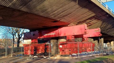 A temporary prop underneath the Gateshead Flyover. It is a huge red metal structure and is fenced off. The flyover above is made from concrete.