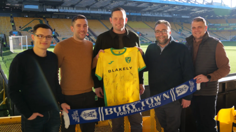 Five men standing inside Norwich City's football stadium holding a Norwich City shirt and holding a blue scarf with Bury Town Football Club written on it