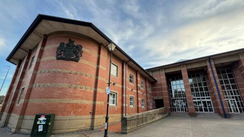 The front of Stafford Crown Court, a brick-built building with a crest and lettering on a rounded part of the building. There are a series of floor-to-roof windows set back behind a series of brick pillars.