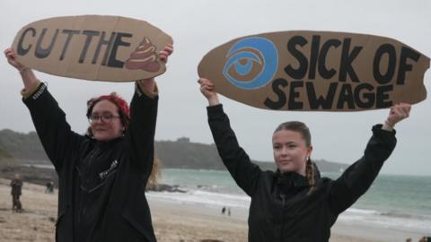 Two Surfers Against Sewage campaigners hold surfboard shaped cardboard signs calling for sewage discharges to be stopped. The two women are stood on a beach on an overcast day.