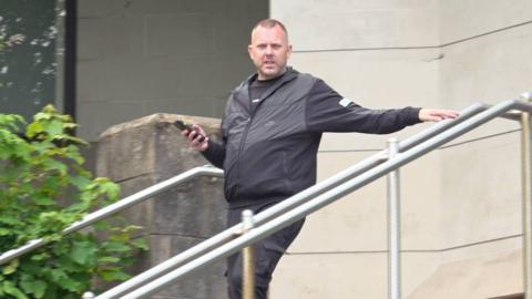 A man in a black jacket, holding a phone and looking towards the camera