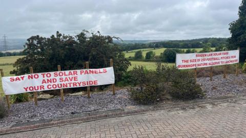 Two banners have been attached to wooden stakes at the top of a hill. The banner on the left reads "Say no to solar panels and save your countryside". The one on the right says "Maiden Law solar farm the size of 250 football pitches from Burnhope nature reserve to Lanchester".