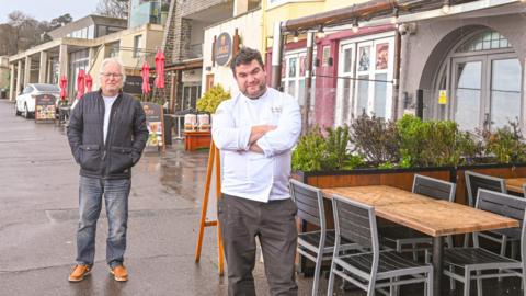 Larry Gibbons and Tom Robinson stand outside their cafes which are next door to each other. Tom is wearing a white chef's jacket and has his arms folded. Larry is wearing black jeans and a jacket and has his hands in his pockets.