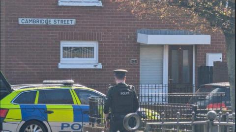 A police officer stands with his back to the camera - in front of him is a police car with battenberg markings. Behind the car is a brown-brick house with a small window, a brown PVC door and another window beside it. A street sign on the house reads "Cambridge Street".