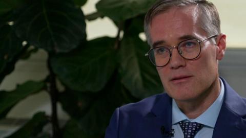 Chris Woodroofe with grey hair and black glasses wearing a blue suit and tie. He is sitting on a leather sofa with a large plant to his left. He looks impassively.