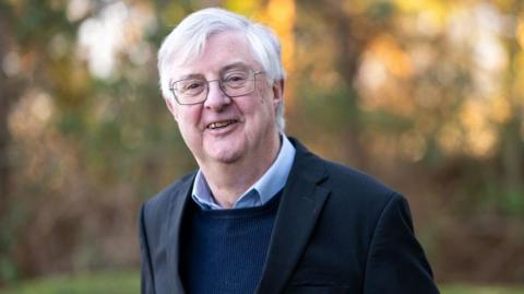Mark Drakeford wearing a dark suit jacket, dark blue jumper and light blue shirt without a tie. His collar is tucked into the jumper.