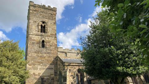 The crenelated tower of St Cuthbert's Church emerging over a tree. The roof of the main body of the church can also be seen.
