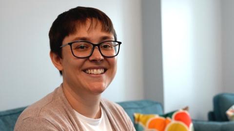 A woman with short brown hair and dark rimmed glasses is sat on her sofa. She's wearing a white top and beige cardigan and is smiling at the camera.