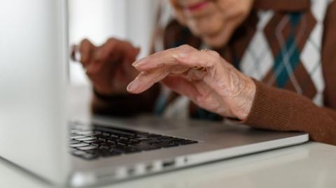 Close‑up of an older person’s hands typing on a laptop keyboard, wearing a brown argyle‑patterned sweater, with the background softly blurred.