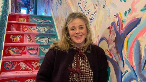 A blonde women smiles in front of a colourful staircase in the hallway of her family home. Painted birds cover the walls.
