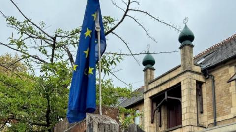 Catherine Bearder, who has short greying, brown hair and is wearing a pale grey, hooded sweater, thin-rimmed round glasses and pearl earrings, pictured outside her home with a Council of Europe flag flying from the top of a tall wooden gatepost. There is also a high brick wall with a tree on the other side