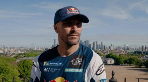 Two-time Dakar Rally Champion Sam Sunderland posing for a picture with his racing outfit against the London skyline. It is a sunny day.