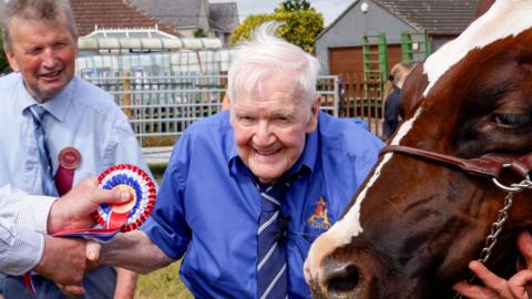 An elderly man with grey hair in a blue shirt and tie with a golden Ayrshire logo on it has a brown cow to his left and is being presented with a red, white and blue rosette. Another man in a lighter blue shirt is looking on behind him