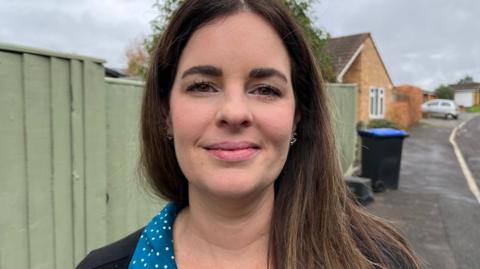 Joeanne looking at the camera smiling. She has long brown hair and is wearing a blue shirt with white spots and a black top. Behind her is a light green fence. 