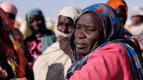A head-and-shoulders image from the side of a woman in a coloured headscarf and pink top. Other women, out of focus, can be seen in the background.